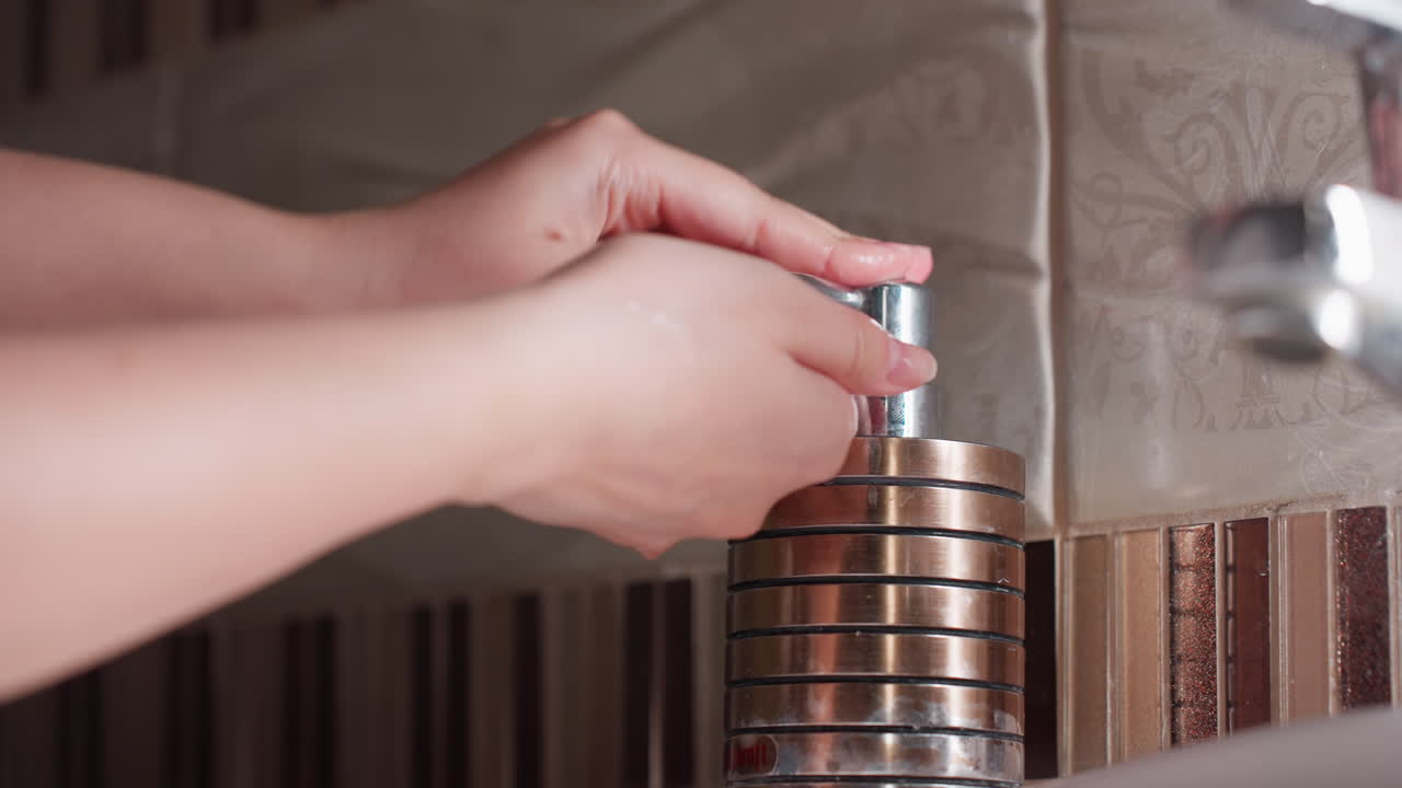 Hand view wet female hands reach toward soap dispenser tip, other hand presses pump, green gel drop forms, chrome bottle on sink beside patterned tiles, hygiene action after wash