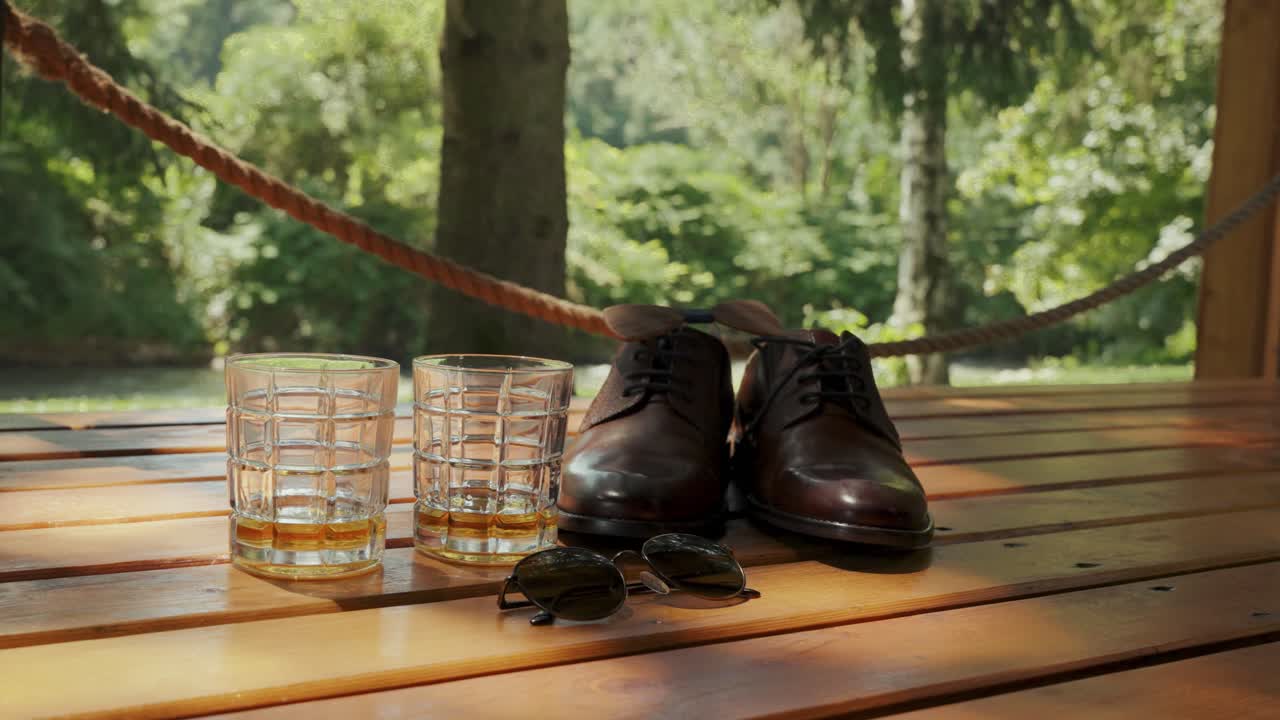 Whiskey glasses, shoes and sunglasses on a table with a garden background.
