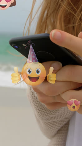 Animación-De-Emoticonos-Sobre-Las-Manos-De-Una-Mujer-Caucásica-Usando-Un-Teléfono-Inteligente-En-La-Playa