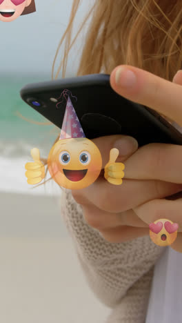 animation of emoticons over hands of caucasian woman using smartphone at beach