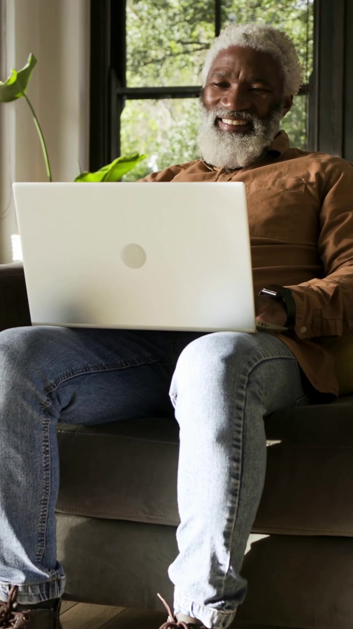 Vertical video: Noticing laptop, man fixing hair, smiling, waving on sofa by window in brown shirt