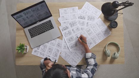 top view zoom out shot of asian male artist talking on smartphone while drawing storyboard for the film on the table with a laptop and headphones in the studio