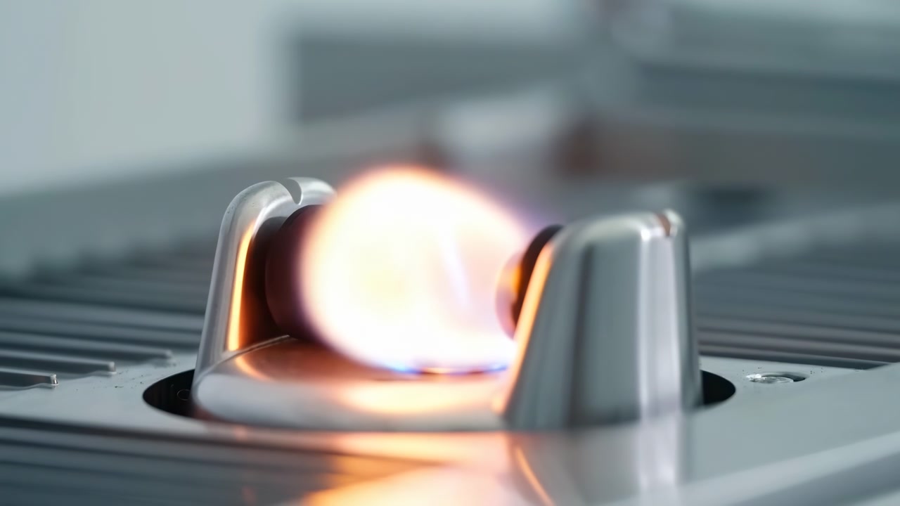 Low-angle close-up shows blue-orange gas flame rising from round burner assembly on ribbed stainless cooktop, with polished chrome support posts, reflective metal surfaces, and strong heat glow