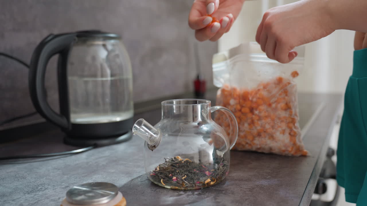 Creating Herbal Tea Infusion Daily, Handplacing Citrus Berries Into Steaming Teapot, Preparing Aromatic Herbal Infusion By Adding Orange Berries Into Glass Teapot In Peaceful Kitchen Setting