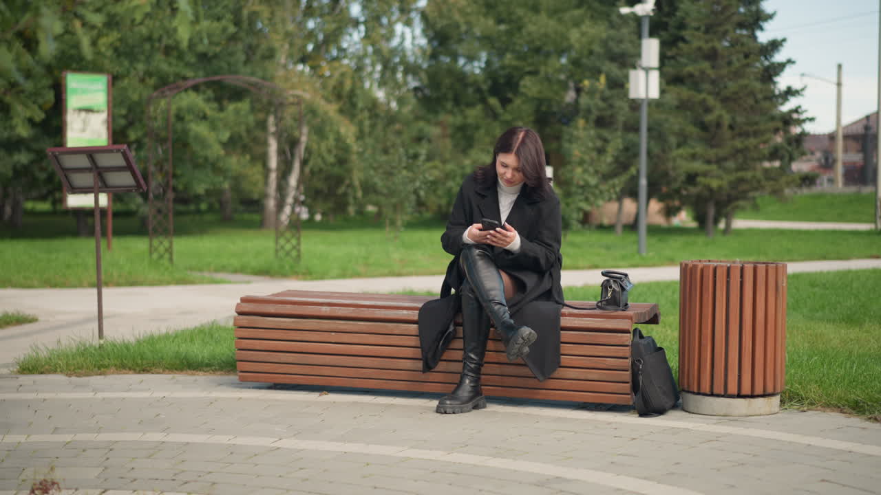 mujer sentada en un banco del parque, usando un teléfono inteligente, vestida con abrigo y botas negras, rodeada de árboles verdes y exuberantes, atmósfera pacífica y relajada en un entorno urbano al aire libre