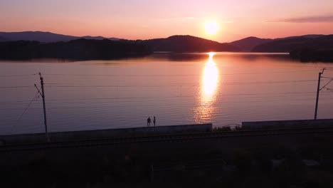 Toma-Aérea-De-Una-Pareja-Caminante,-Vía-Férrea,-Cielo-Despejado,-Puesta-De-Sol-Y-Un-Enorme-Lago-Naranja