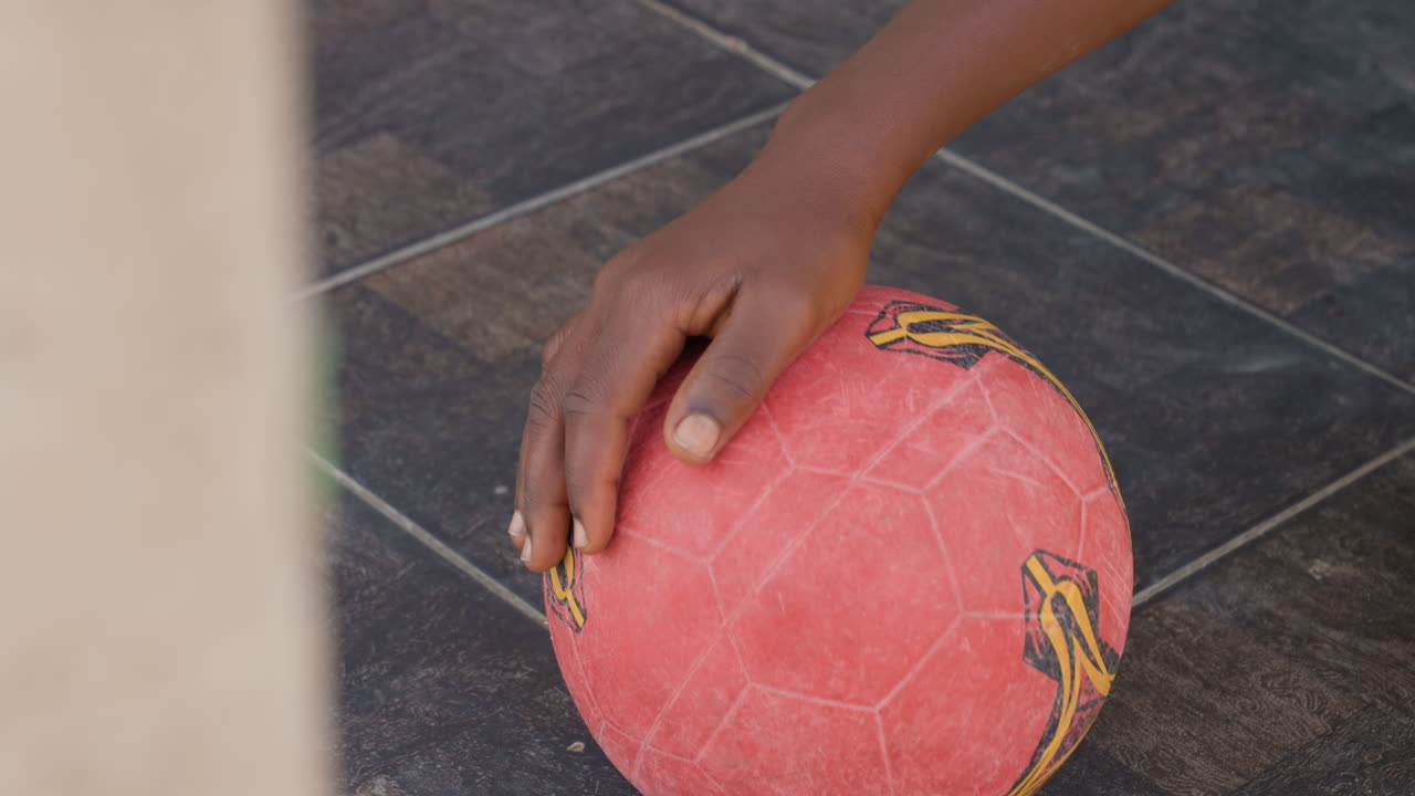 Mão colocando bola de futebol vermelha no azulejo, palma de jovem negro ajeitando a bola em quadra de azulejos, foco e calma pré-jogo, preparação urbana autêntica para partida de rua com superfície texturizada e desgastada.