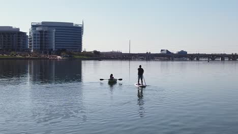 Slow-motion-of-a-male-standup-paddleboarder-and-female-kayaker-on-Tempe-Town-Lake,-Tempe,-Arizona