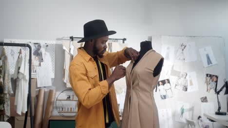 stylish young man clothing designer wearing hat making marks with pins on dress in mannequin in the studio