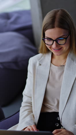 pleasant-looking woman in glasses leans back and sits comfortably in bean bag chair with laptop on her legs. modern office work concept. vertical video