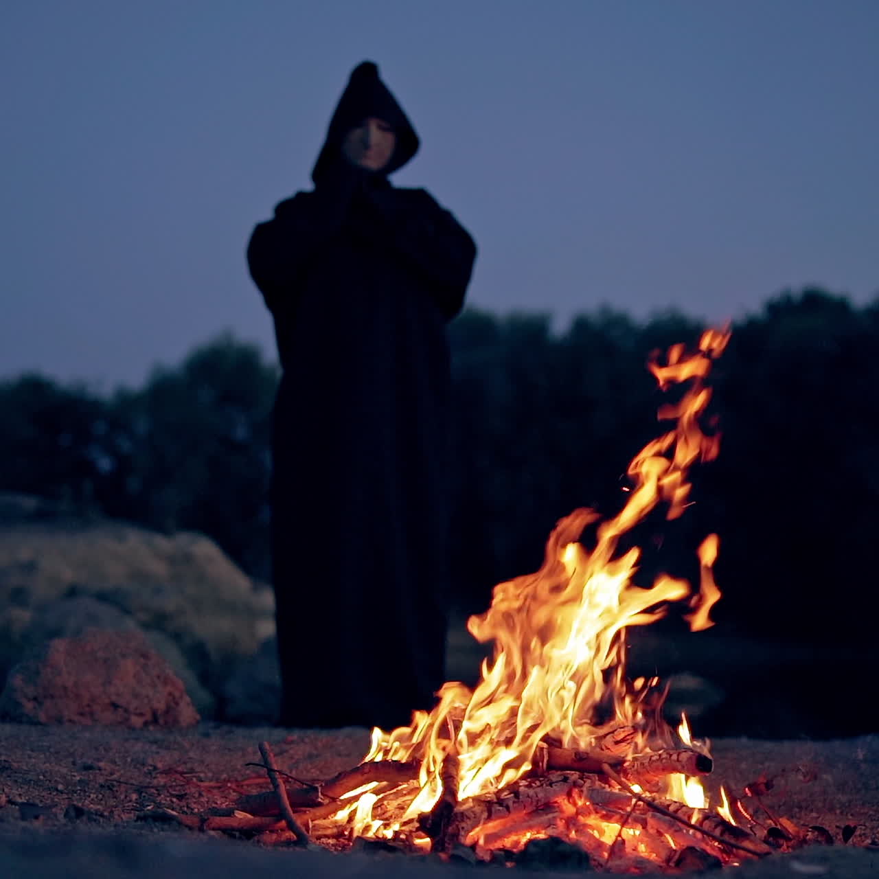 Halloween. Powerful witch or wizard in black costume stands and watches the fire flame in the evening.