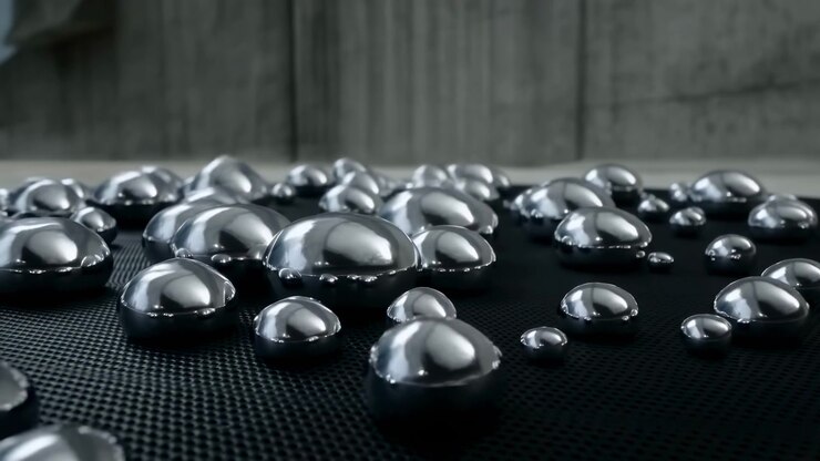 Array of chrome-like spherical droplets resting on dark rubber or polymer mat with microgrid texture in blurred concrete-walled interior, camera movement or rack focus gradually brings midground and