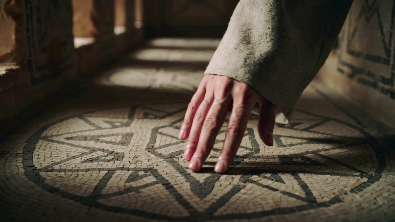 Entrando da destra, una mano ammantata scende, toccando il pavimento a mosaico e tracciando un motivo a stella, con spazio per il testo.