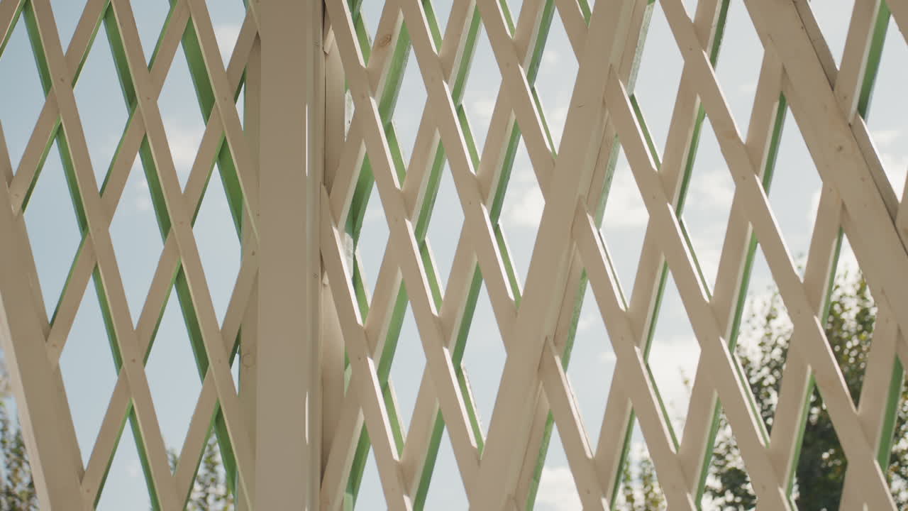 Allestimento soleggiato in giardino. Padiglione in legno con accenti verdi. Gazebo esterno con calde tonalità di legno e ombre tenui. Struttura reticolata illuminata dal sole con travi in legno e atmosfera da giardino