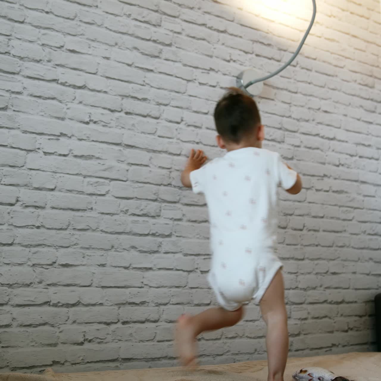 Seorang anak laki-laki balita mengenakan romper berlari ke sakelar lampu. Anak itu menyalakan lampu sambil tersenyum dan melompat dengan riang
