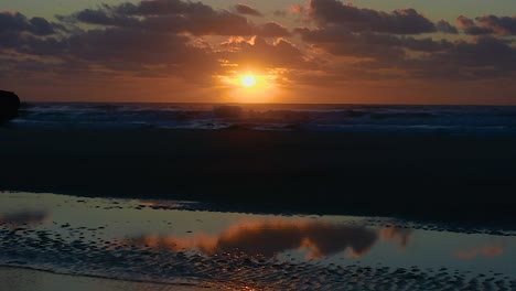 Dramatic-view-across-a-deserted-beach-with-the-sky-reflected-in-pools-of-water-and-a-stunning-golden-sunrise-or-sunset-with-a-silhouetted-4x4-truck-driving-past-with-fishing-gear