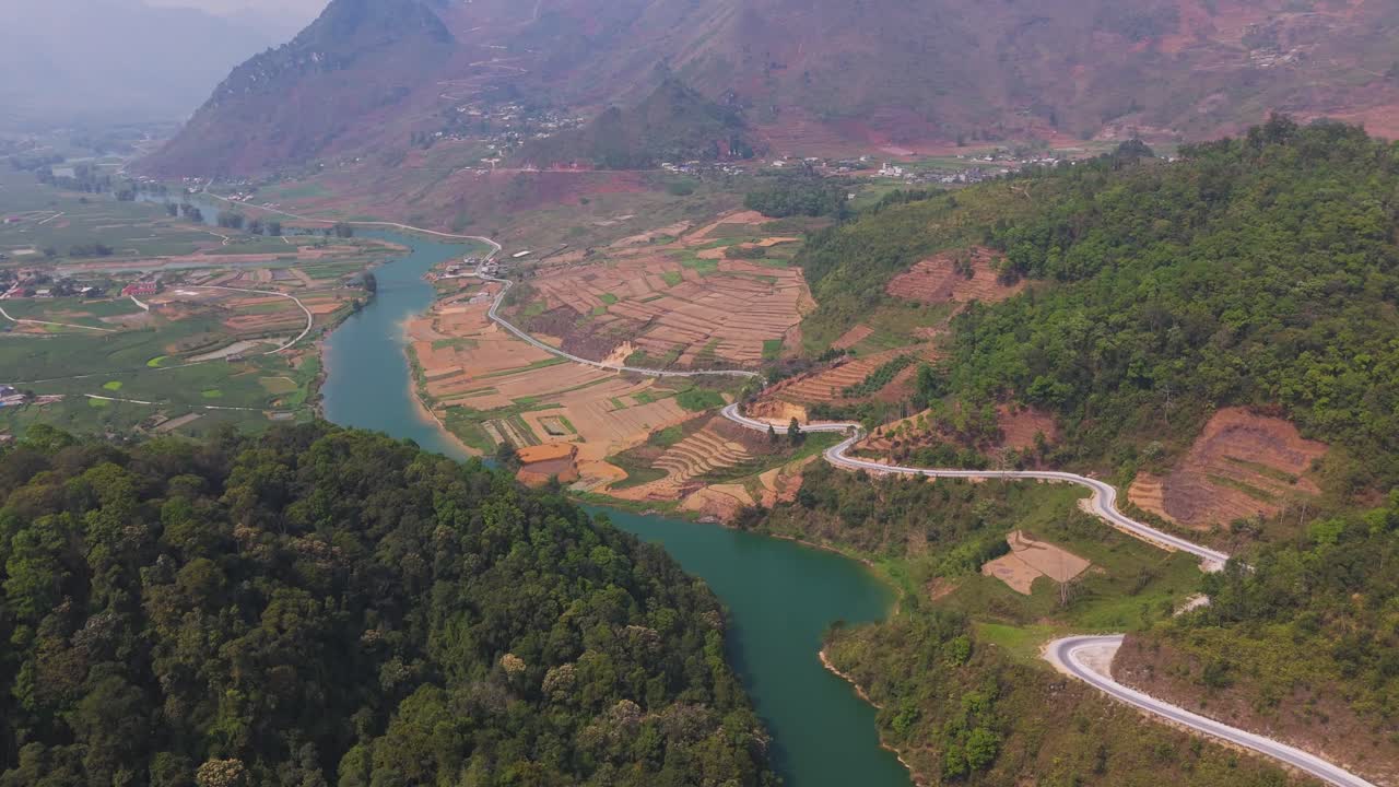 Fly above Ha Giang’s scenic serpentine roads with motorcyclists weaving between a river and rugged mountains.
