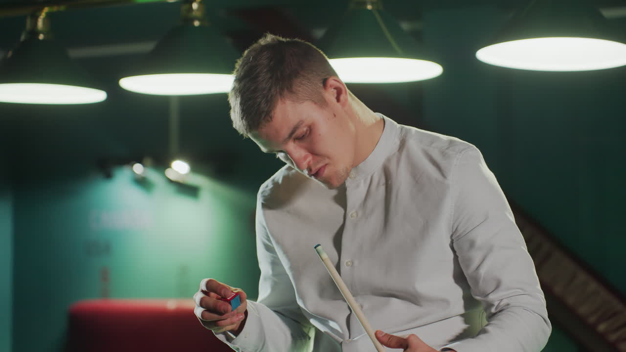 색 셔츠를 입은 게임맨이 큐 스틱을 들고 조심스럽게 은 개를 적용하여 희미하게 조명된 빌리어드 홀에서 집중된 표정을 표시합니다.