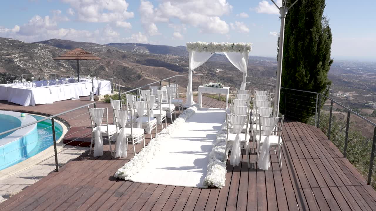 Delle sedie sono disposte lungo una navata bianca per una cerimonia nuziale. Dei fiori costeggiano il percorso. Una piscina e dei tavoli sono visibili in una zona panoramica. Il sole splende sulle colline.