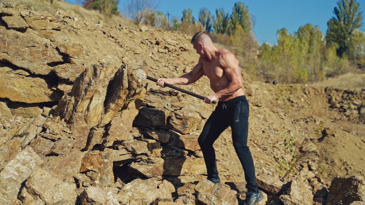 Shirtless bodybuilder splitting rocks with a sledgehammer. Strong man breaking stones with a steel hammer in the mountains. Slow motion.