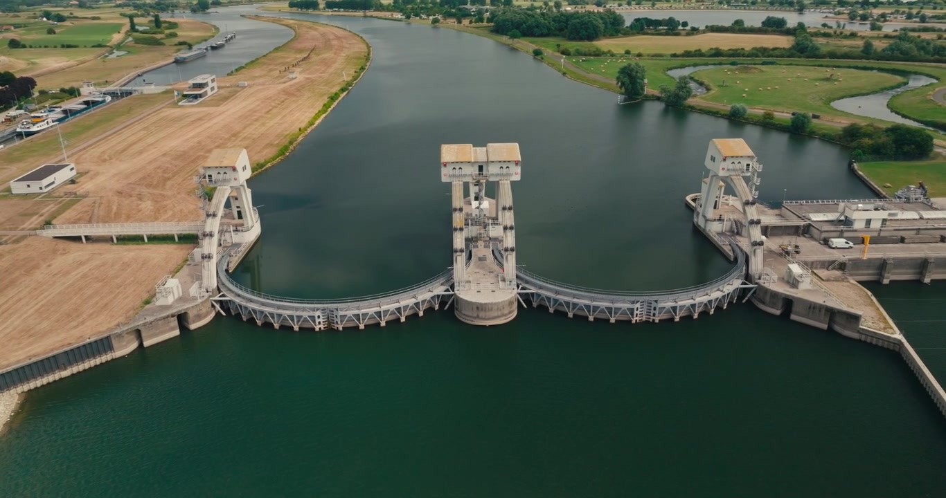 Drone orbits a hydroelectric power station in a Dutch canal, revealing renewable energy infrastructure surrounded by peaceful countryside, farmland and waterways on a sunny day.
