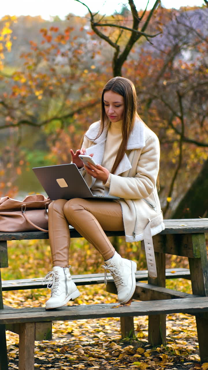 Beautiful brunette sitting on the wooden table with laptop on her knees. Woman looking at the phone in her hands. Autumn nature at backdrop. Vertical video