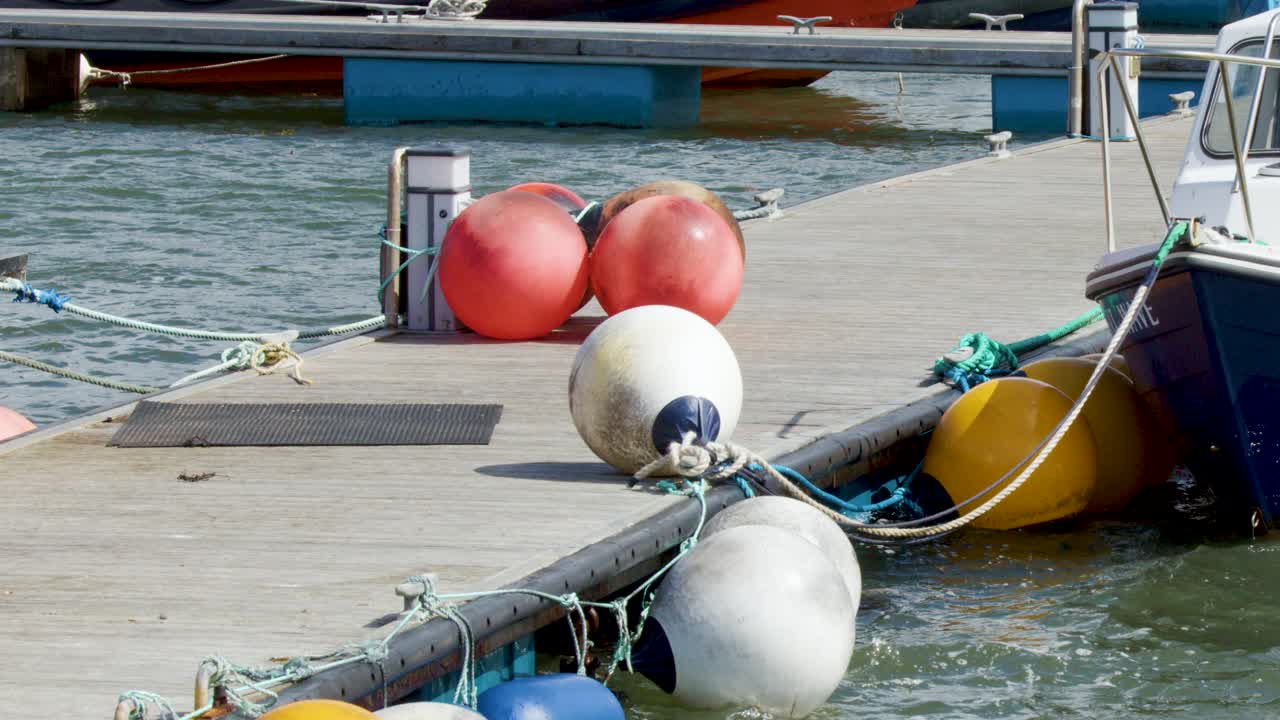 Colorful buoys gently move in the water beside moored boats at a sunlit Cromarty harbour dock, captured in a steady, wide shot