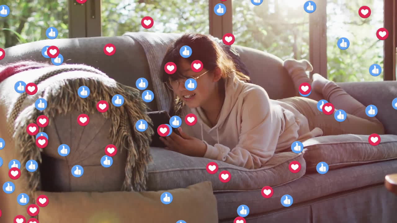 Animation of social media icons over asian woman using tablet on sofa