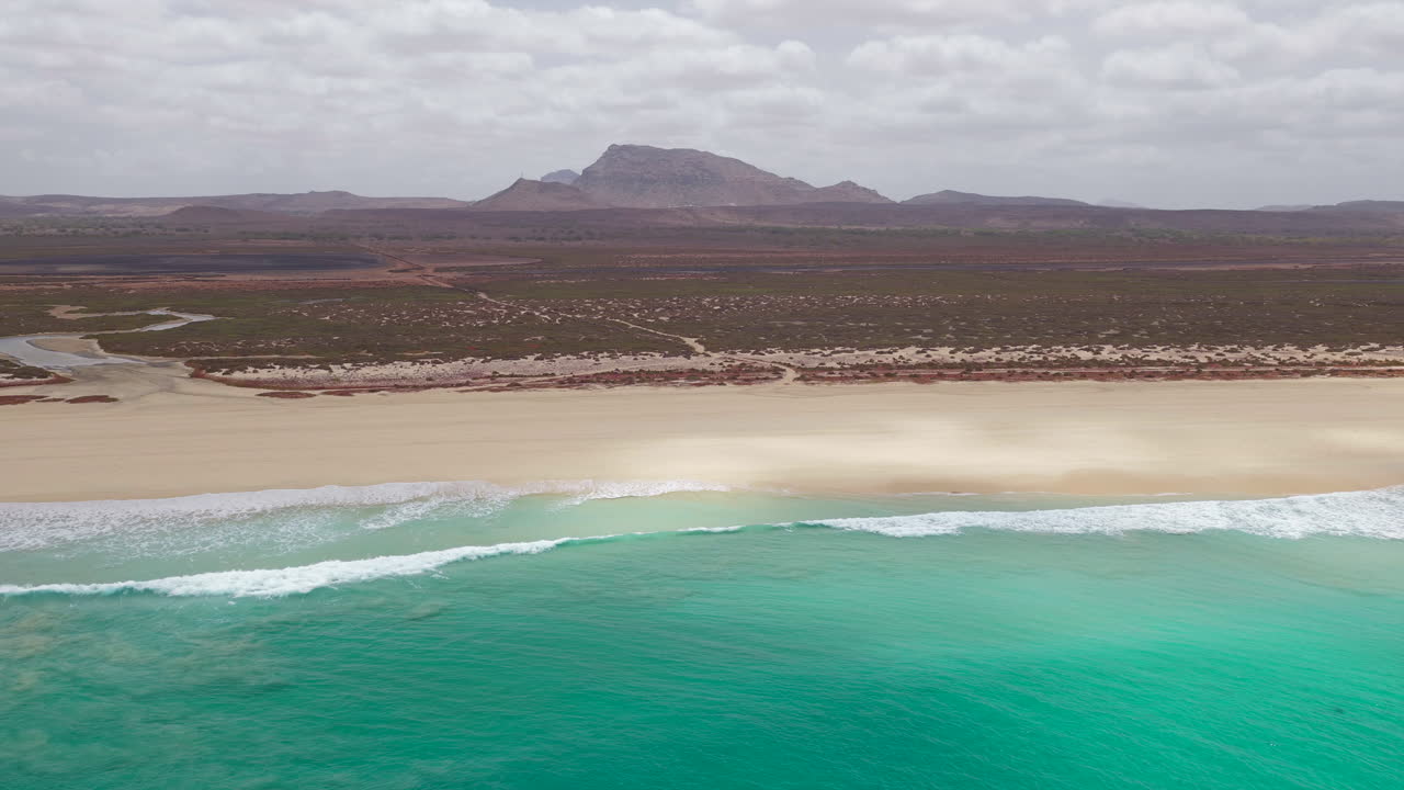 Drone footage of Santa Monica Beach,showcasing endless white sand, turquoise waves, and a pristine,background volcanic mountain and desert,Boa Vista, Cape Verde