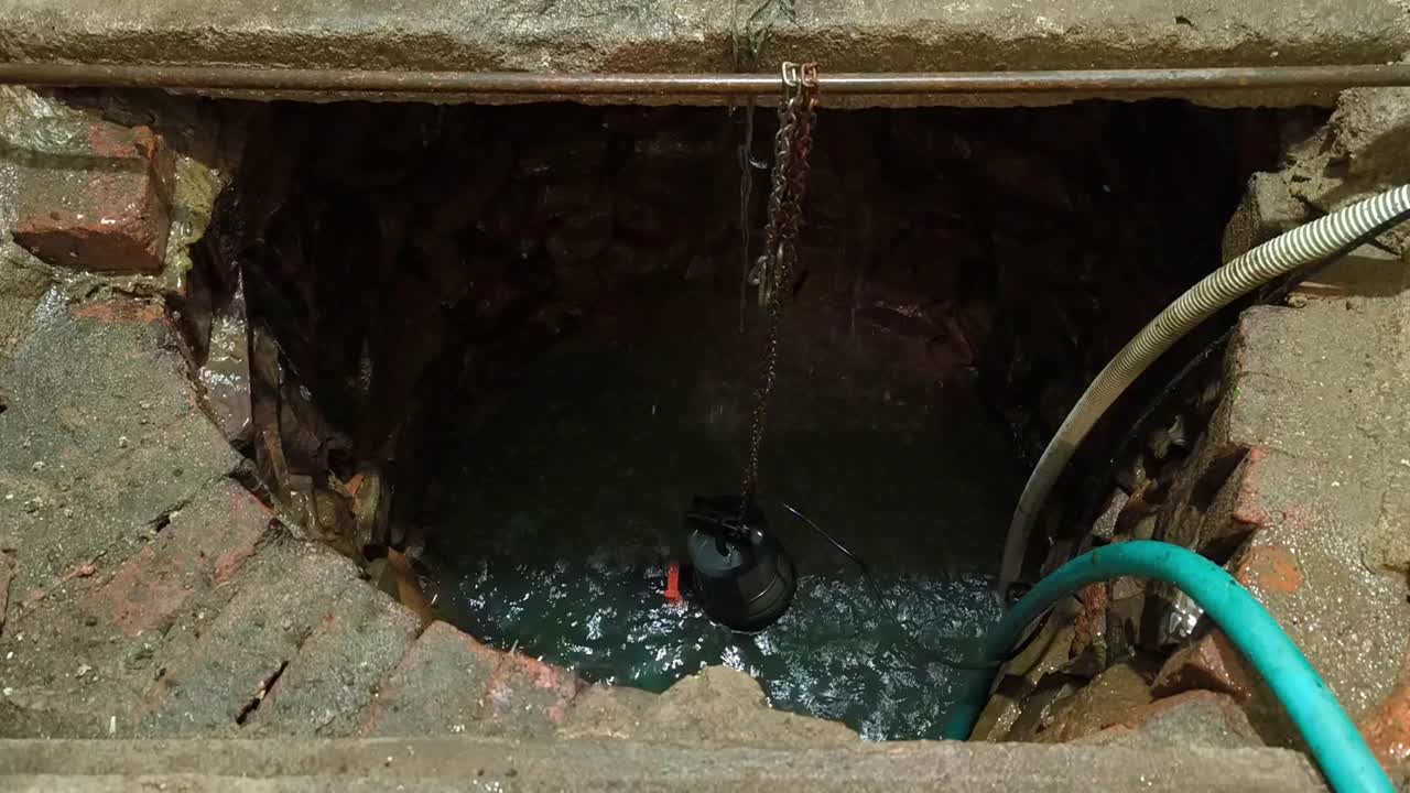 Vista ravvicinata di un pozzo allagato che viene drenato da una pompa durante condizioni di piena, con forte afflusso di acqua fangosa all'interno di una vecchia struttura in muratura, gestione pratica delle acque sotterranee e rimozione di emergenza dell'acqua.