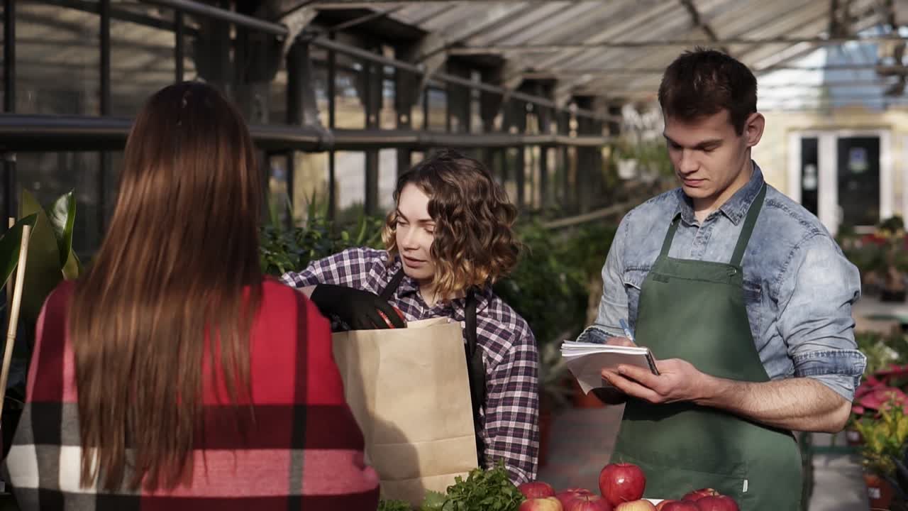 la vendedora europea con delantal está dando comida orgánica al cliente en el invernadero. la mujer está empaquetando verduras, frutas y verduras en una bolsa de papel marrón mientras el hombre toma notas. la gente y el concepto de estilo de vida saludable