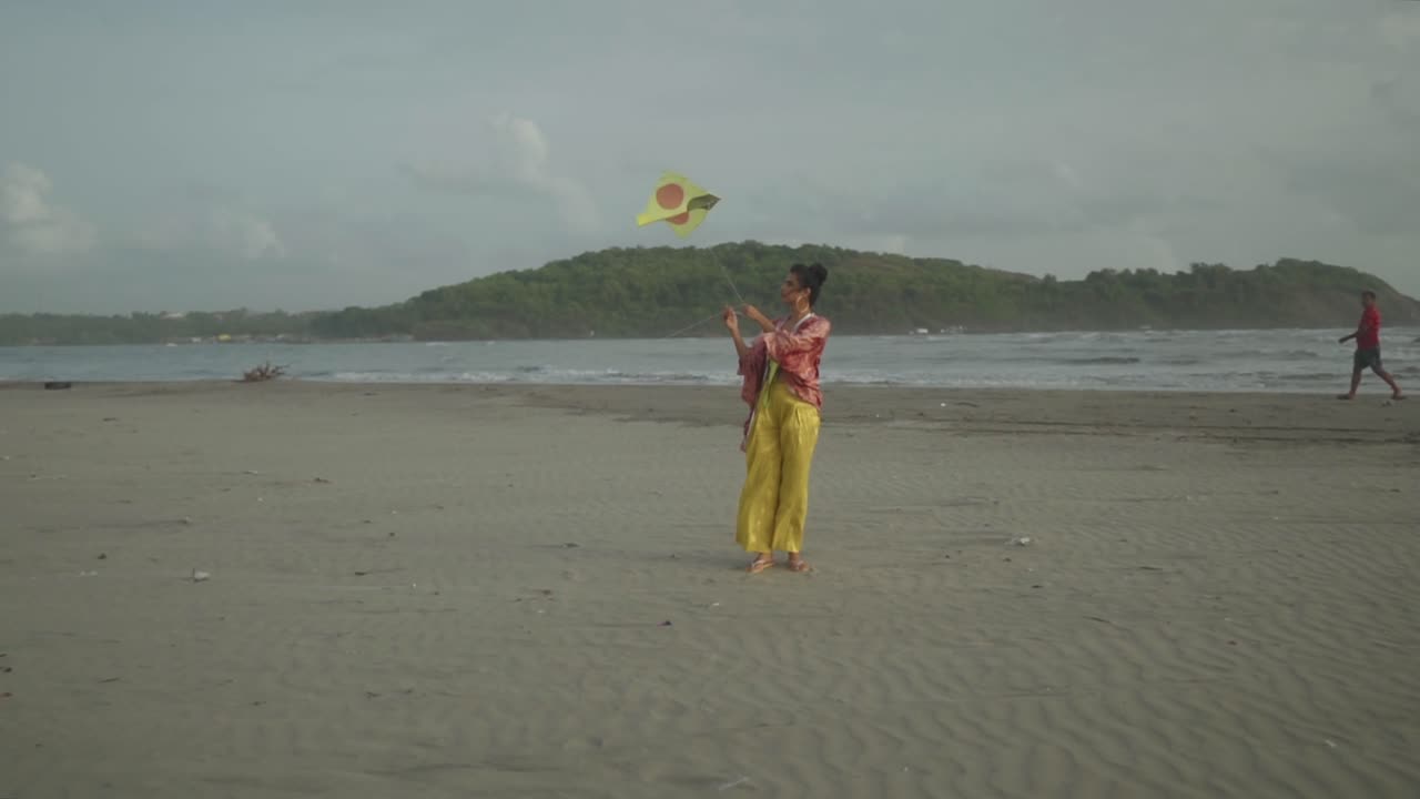 A young Asian model adorned in yellow outfit and falling her kite joyfully while standing on a beachside location