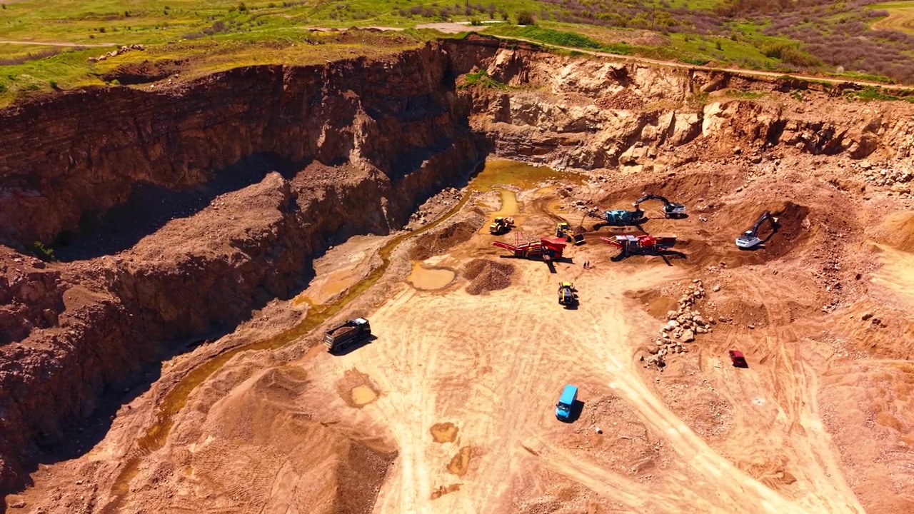 Deep open pit rock quarry with heavy machinery and cliff walls. High perspective reveals crushing equipment and vehicles inside a large brown rock pit surrounded by green grass.
