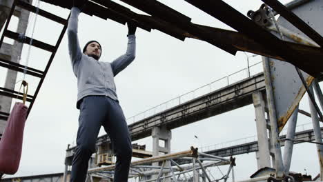 joven deportista guapo haciendo pull-ups en la construcción de metal en las ruinas de una fábrica abandonada en una mañana nublada