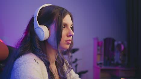 side view of woman wearing white headphones and fluffy sweater seated in cozy room, concentrating on gaming with relaxed expression while eating popcorn