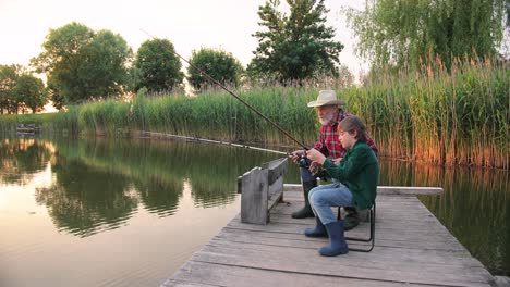 side view of a teen boy sitting with his grandfather on the lake pier, talking and fishing together