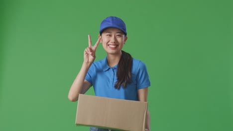 푸른 유니폼을 입은 아시아 여성 택배기사는 스튜디오에서 녹색 스크린 배경에 상자를 배달하는 동안 몸짓 평화와 미소를 짓고 있습니다.