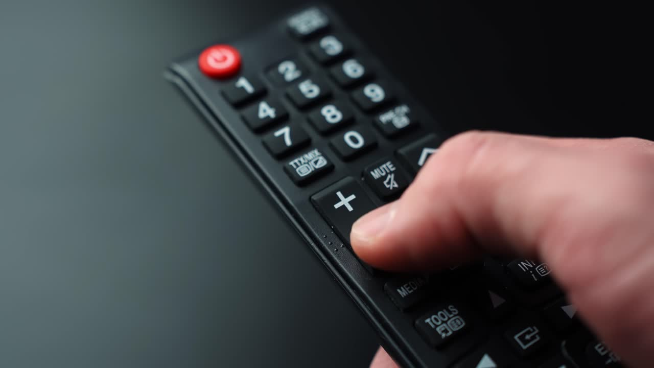 Primo piano di un pollice che abbassa il volume su un telecomando TV su uno sfondo scuro con bassa profondità di campo e spazio per il testo, ideale per streaming, intrattenimento domestico e regolazione audio.