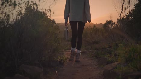 Front-view-of-a-woman-walking-in-a-hike-during-a-sunset-or-sunrise-and-holding-a-flask-bottle,-slow-motion-close-up-shot