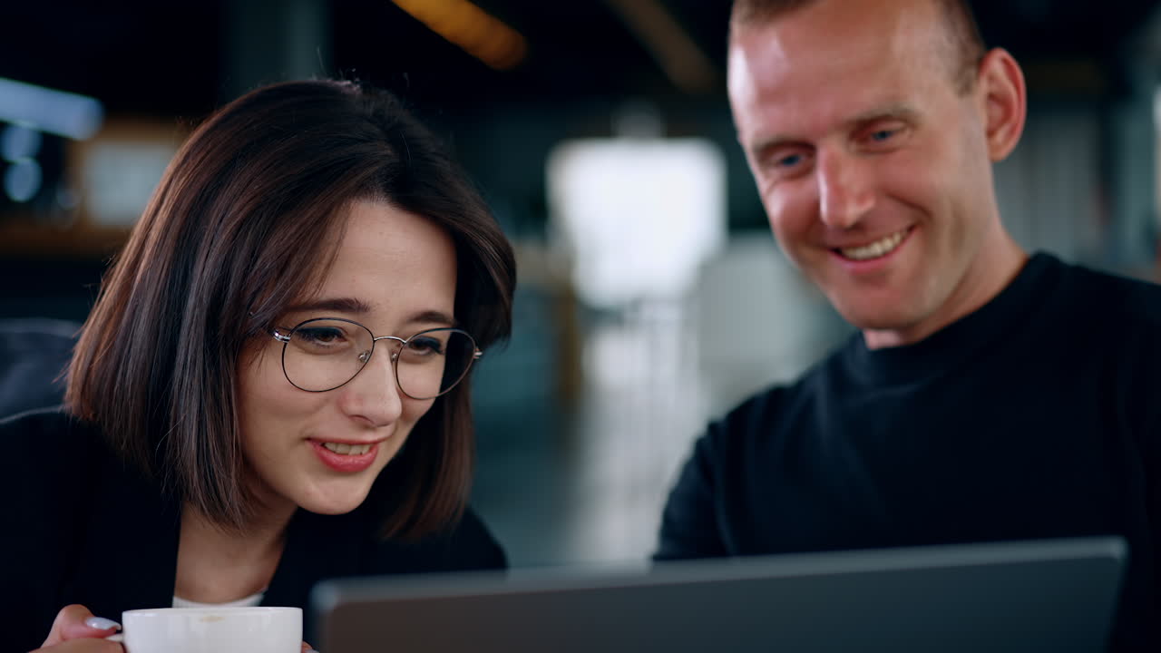 Man and woman look at laptop screen laughing. Lady wearing glasses holds a cup in hand. Close up.