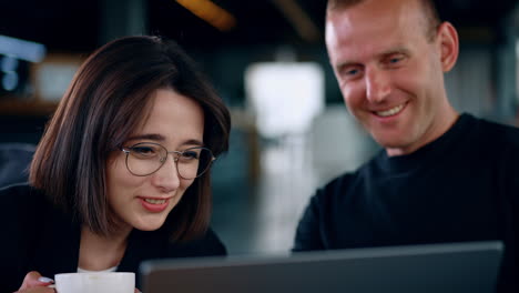man and woman look at laptop screen laughing. lady wearing glasses holds a cup in hand. close up.
