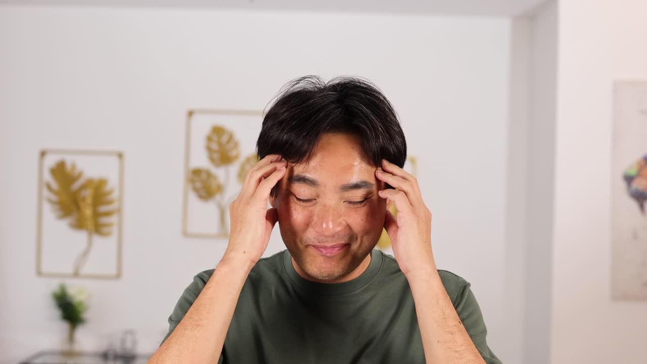 An Asian man in a green shirt sits indoors, touching his temples with both hands, displaying a headache gesture under soft, natural lighting. Camera remains steady