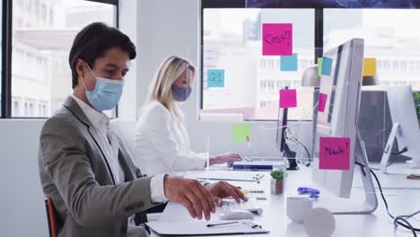 Diverse-business-colleagues-wearing-face-masks-working-at-desks-in-office-with-hygiene-screen-betwee