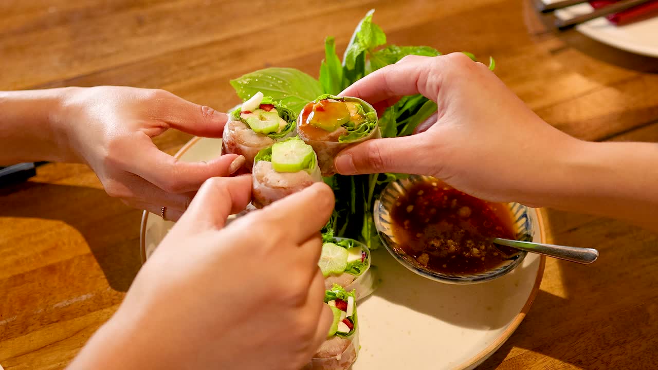 Hands preparing and dipping Vietnamese pork rolls in sauce at a Bangkok restaurant. Bright lighting enhances the vibrant, fresh ingredients