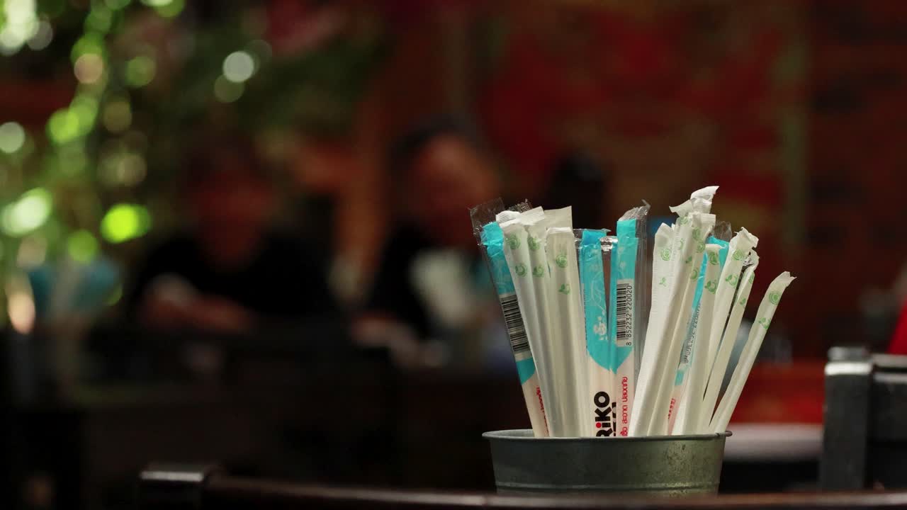 A bustling restaurant in Phuket, Thailand, with straws prominently displayed in the foreground and blurred patrons in the background