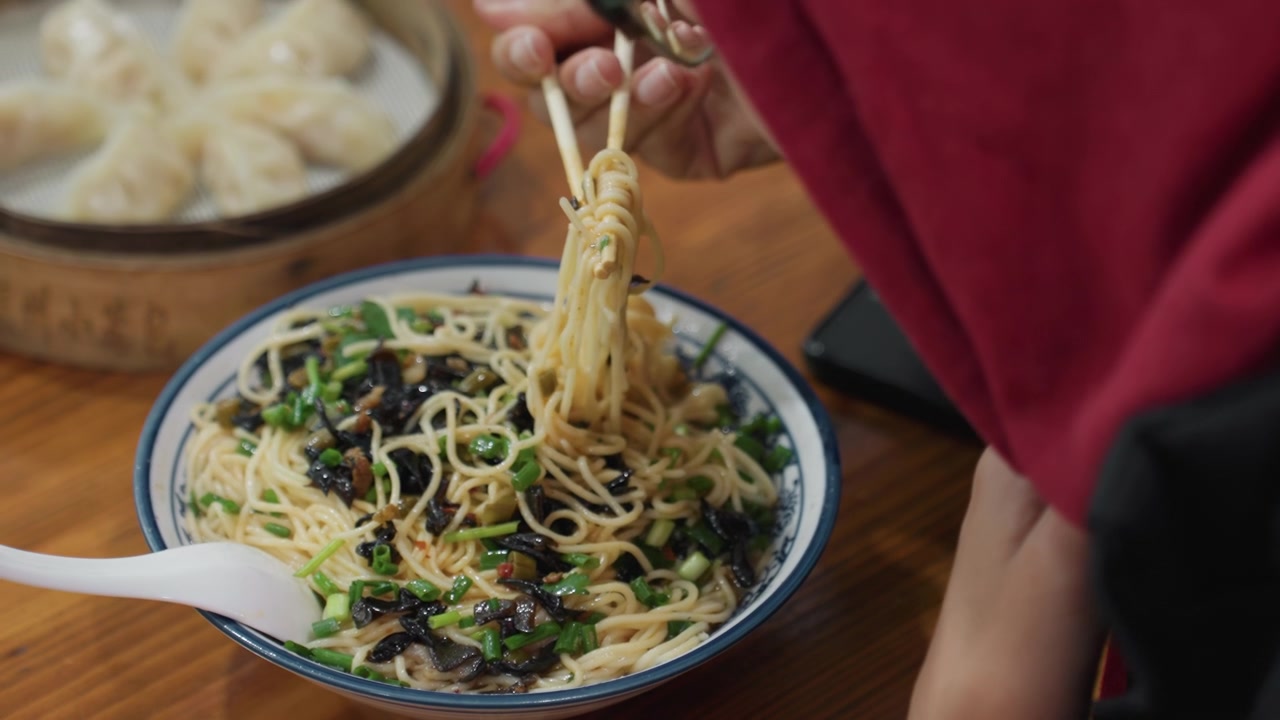 Mani che sollevano spaghetti accanto a un cesto di ravioli, bacchette che prendono fili sopra una ciotola di ceramica, vaporiera di bambù con ravioli sullo sfondo, smartphone su tavolo di legno, manica di giacca casual e saporito