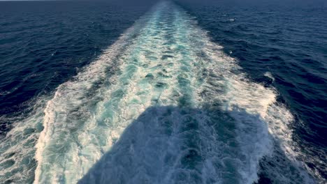 rear view of wide water wake left from cruise ship on sea water surface with horizon in background and shadow of people leaning over parapet of deck