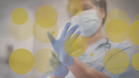 animation of yellow spots over caucasian female doctor in face mask putting gloves on