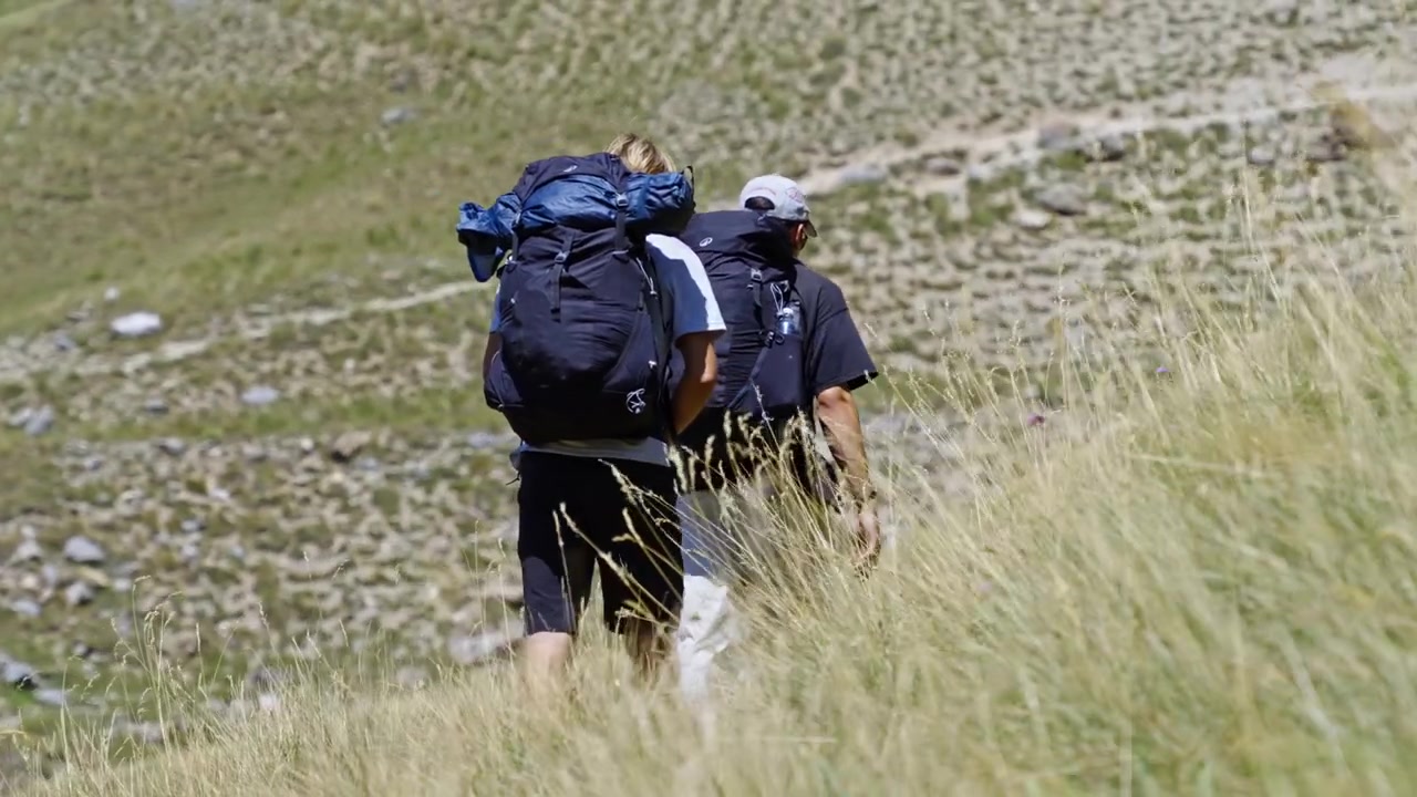 To ryggsekkturister går langs en smal fjellsti gjennom høyt, tørt gress på en solrik sommerdag