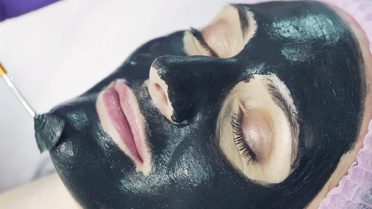 Procedure for applying a black mask to the face of a beautiful woman in the beauty salon. Cosmetologist is putting purifying mask with a brush. Close-up